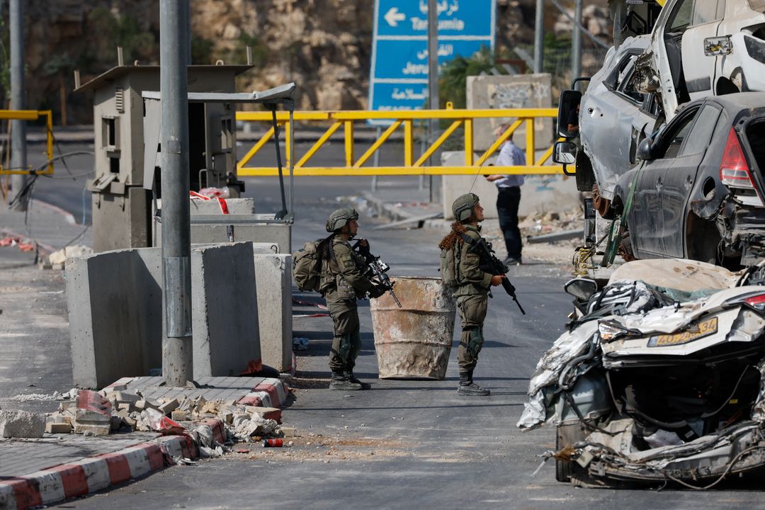 Two Israeli soldiers holding rifles stand at a checkpoint by concrete barriers and a yellow gate, beside a stack of crushed cars.