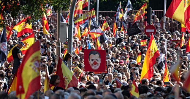 A dense crowd waves Spanish flags on a Madrid street; in the middle, a placard shows a man’s face with a red clown nose.