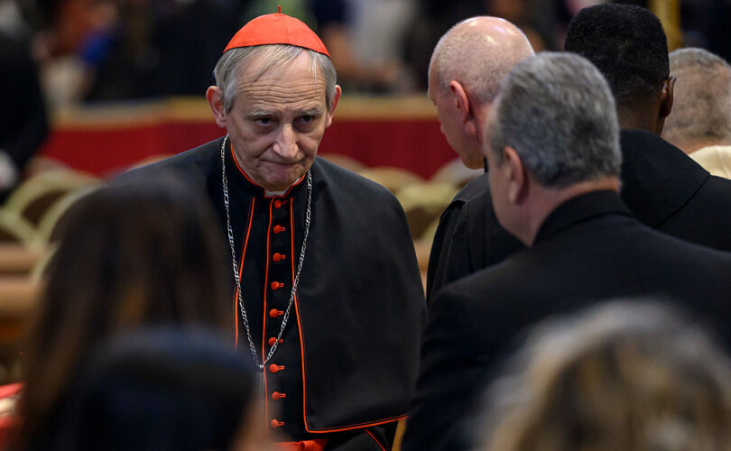 Cardinal Matteo Zuppi, in a red skullcap and black cassock with red buttons, walks past clergy in a crowded hall, looking toward the camera.