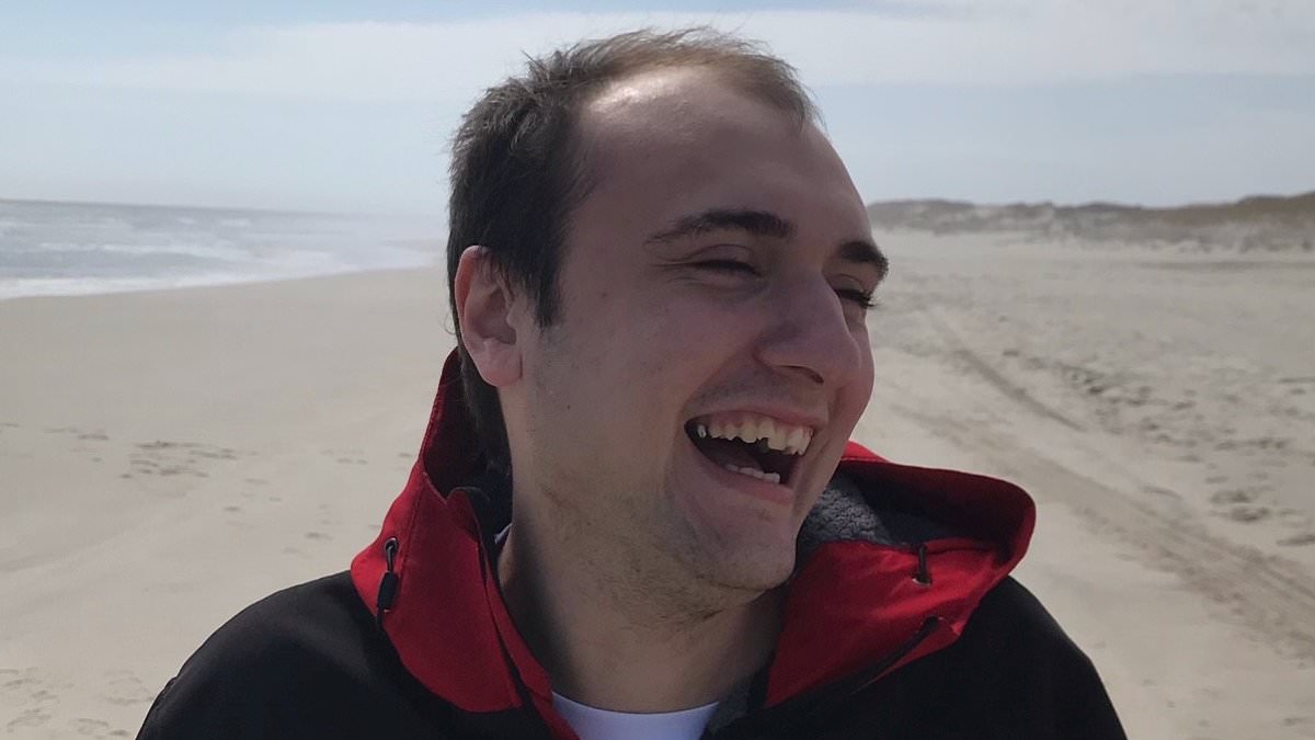 A young man in a black jacket with a red hood lining laughs on a sandy beach under a pale sky.
