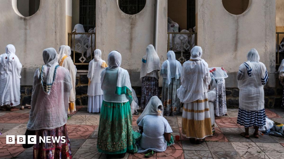 Women in white shawls stand and kneel in prayer facing a church wall, seen from behind, on a tiled courtyard.